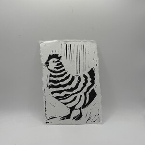 May include: Black and white linocut print of a hen. The artwork features a stylized hen with striped body and textured details. The print is on a rectangular piece of textured, off-white paper with a deckled edge.