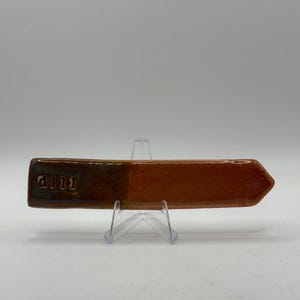 以下が含まれることがあります： 茶色と緑色の釉薬を施した、手作りのセラミック製の矢印型の標識。標識の左側には「d111」の文字が入っています。矢印は透明なプラスチック製のスタンドに展示されています。