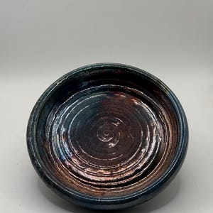 May include: A round ceramic bowl with a dark, iridescent glaze. The interior features visible concentric circular ridges and a metallic, copper-toned finish that shifts to deep blue and black hues along the thick, rounded rim of the vessel.