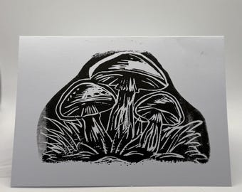 May include: A black and white linocut print featuring three mushrooms with detailed caps and stems, set against a backdrop of stylized grass. The print is on a rectangular, light gray card.