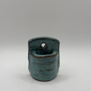 May include: A small, wall-mounted ceramic planter with a glossy teal glaze and a dark brown rim. The vessel features a rounded, cup-like shape with a flat back panel containing a single circular hole for hanging on a wall or vertical surface.