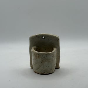 May include: A small, off-white ceramic wall pocket with a speckled finish. The piece features a flat back panel with a single circular hole at the top for mounting and a rounded, protruding cup-shaped container attached to the front surface.