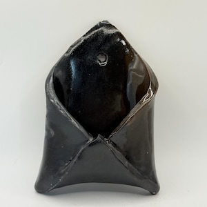 May include: A wall-mounted ceramic pocket with a glossy black finish. The object has a pointed top section featuring a small circular hole for hanging, and a wide, open-top envelope-like shape with curved edges and a smooth, reflective surface.