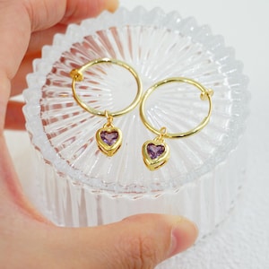 May include: A pair of gold hoop earrings with heart-shaped purple gemstones. The earrings are displayed on a clear, textured glass surface. The earrings are held by a hand.
