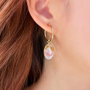 May include: Gold hoop earring with a clear, oval-shaped charm. The charm contains pink and gold floral elements. The earring is worn on a person's ear, with reddish-brown hair visible in the background.