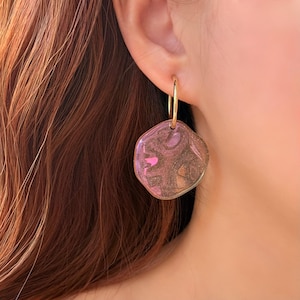 May include: Gold hoop earrings with a unique, irregular-shaped pendant. The pendant features a blend of pink, gold, and brown hues, creating a shimmering, iridescent effect. The earrings are shown on a person's ear, highlighting the jewelry's design and color.