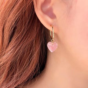 May include: A gold hoop earring with a pink heart-shaped charm. The heart charm has a marbled appearance. The earring is shown on a person's ear.