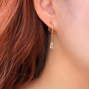 May include: A gold hoop earring with a dangling rectangular clear gemstone. The earring is attached to a chain that hangs from the hoop. The earring is worn on a person's ear.