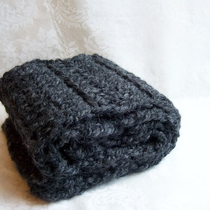 May include: A dark gray knitted blanket folded on a white surface. The blanket is made of a thick, soft yarn and has a cable knit pattern.