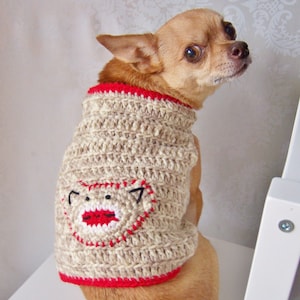 Peut inclure: Un petit chihuahua brun portant un pull en crochet beige pour chien avec une bordure rouge et un motif de visage de singe aux lèvres rouges.