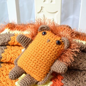 May include: A crocheted orange and brown lion plush toy with a fluffy mane, sitting on a multi-colored crocheted blanket.
