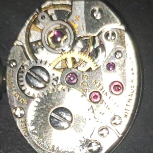 May include: Close-up of a vintage watch movement with intricate gears, jewels, and screws. The silver-toned mechanism features the text "Swiss" and "Wittnauer". The watch movement is oval-shaped.