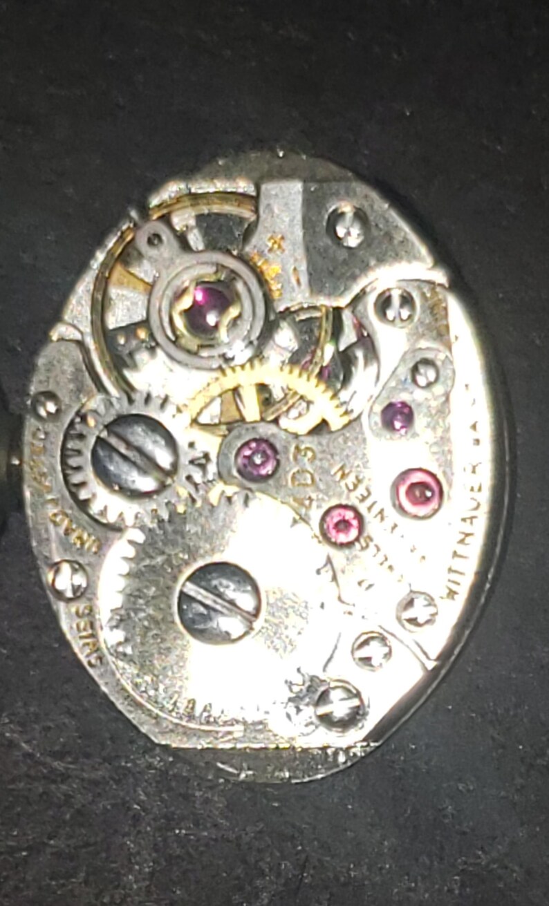 May include: Close-up of a vintage watch movement with intricate gears, jewels, and screws. The silver-toned mechanism features the text "Swiss" and "Wittnauer". The watch movement is oval-shaped.