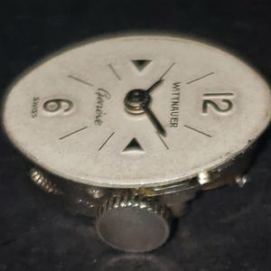 May include: A vintage Wittnauer watch face with a silver-colored dial. The face features black hands, the number 12, and the number 6. The words "Geneve" and "Swiss" are also visible. The watch has a silver-colored crown.