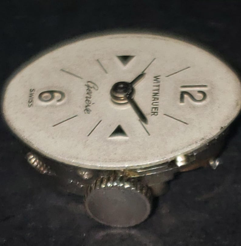 May include: A vintage Wittnauer watch face with a silver-colored dial. The face features black hands, the number 12, and the number 6. The words "Geneve" and "Swiss" are also visible. The watch has a silver-colored crown.