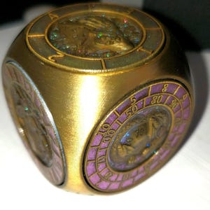May include: A gold-colored, cube-shaped object with circular insets. Each inset features a detailed design, including a central image and surrounding numbers. The cube's sides have a metallic sheen, and the insets are accented with purple and gold.