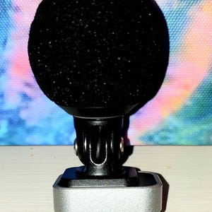 May include: A black microphone with a rounded, textured head, mounted on a black and silver base. The base has a silver rectangular body with a connector at the bottom. The background is a colorful, abstract design.