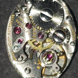 May include: Close-up of a vintage watch movement. The silver-toned mechanism features intricate gears, ruby jewels, and the text "Wittnauer Watch". The watch is marked "Swiss" and "Unadjusted".