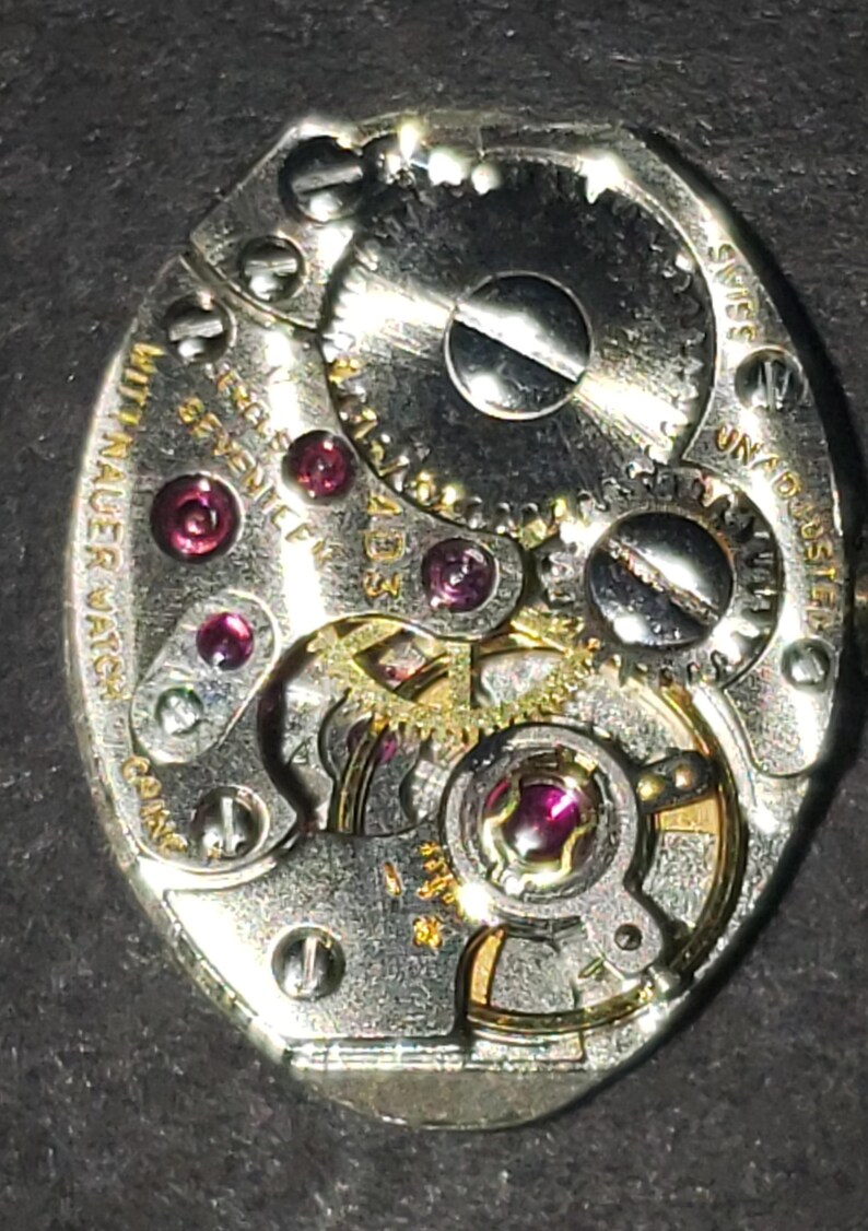 May include: Close-up of a vintage watch movement. The silver-toned mechanism features intricate gears, ruby jewels, and the text "Wittnauer Watch". The watch is marked "Swiss" and "Unadjusted".