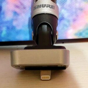 May include: A silver and black Shure microphone with a lightning connector. The microphone has a silver mesh head and a black body. The base is silver and rectangular. The Shure logo is visible on the microphone.