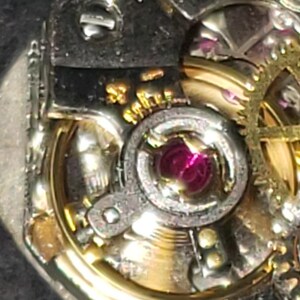 May include: Close-up of a watch mechanism with intricate details. The image showcases silver and gold-toned gears, a ruby jewel, and small screws. The watch parts are arranged in a circular pattern, highlighting the precision of the craftsmanship.