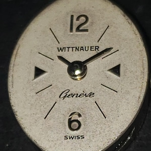 May include: Vintage Wittnauer watch face with a silver-tone oval dial. Features black hands, hour markers, and the numbers 12 and 6. The words "WITTNAUER", "Gen&egrave;ve", and "SWISS" are printed on the dial.