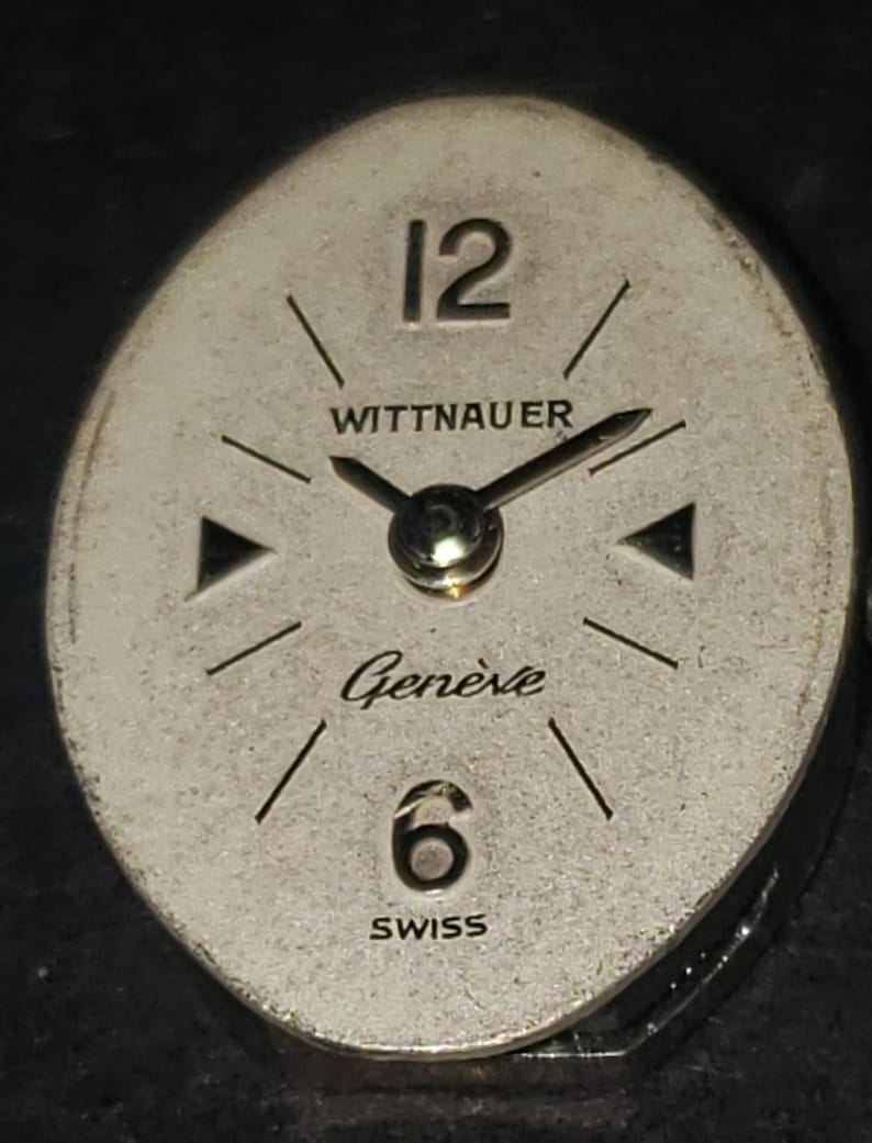 May include: Vintage Wittnauer watch face with a silver-tone oval dial. Features black hands, hour markers, and the numbers 12 and 6. The words "WITTNAUER", "Gen&egrave;ve", and "SWISS" are printed on the dial.