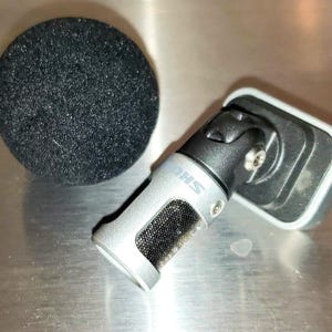 May include: A silver and black microphone with a black foam windscreen. The microphone has a mesh screen and the brand name "SHURE" is visible. The microphone is angled and attached to a black mount.
