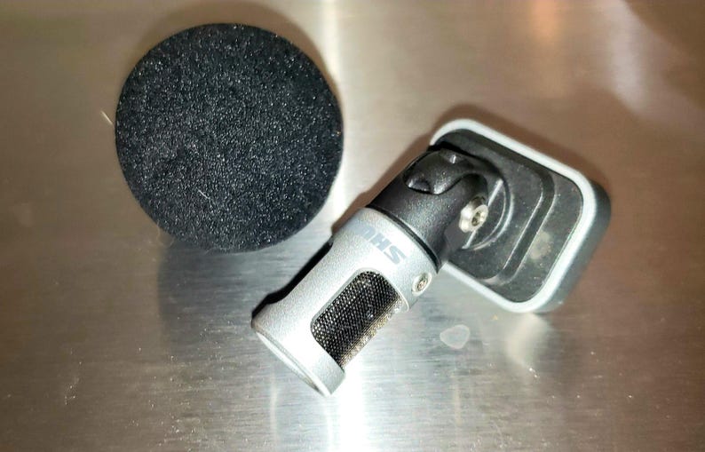 May include: A silver and black microphone with a black foam windscreen. The microphone has a mesh screen and the brand name "SHURE" is visible. The microphone is angled and attached to a black mount.