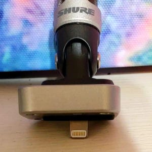 May include: A silver and black Shure microphone with a lightning connector. The microphone has a silver mesh head and a black body. The base is silver and rectangular. The Shure logo is visible on the microphone.