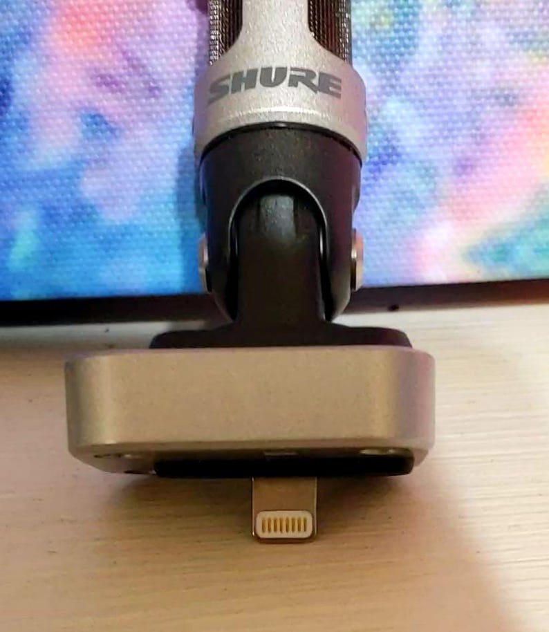 May include: A silver and black Shure microphone with a lightning connector. The microphone has a silver mesh head and a black body. The base is silver and rectangular. The Shure logo is visible on the microphone.