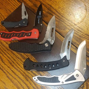 Puede incluir: Una variedad de navajas con diversos diseños de mango y formas de hoja. Las navajas tienen mangos negros, rojos y plateados. Una navaja tiene la inscripción "FIRE FIGHTER" en la hoja.