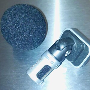 May include: A silver microphone with a black foam windscreen. The microphone has a rectangular base and a pivoting head. The windscreen is a dark gray, circular shape. The microphone is designed for recording.