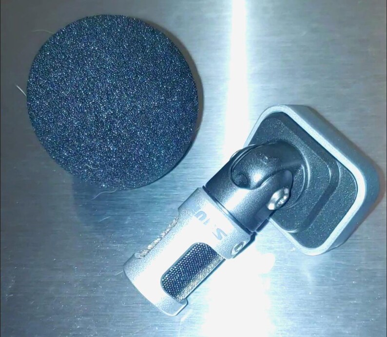 May include: A silver microphone with a black foam windscreen. The microphone has a rectangular base and a pivoting head. The windscreen is a dark gray, circular shape. The microphone is designed for recording.