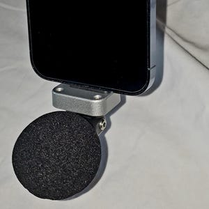 May include: A black smartphone attached to a silver metal bracket with a black, round, foam microphone. The phone is positioned above the microphone, which is connected to the bracket via a small hinge. The background is a light, neutral color.