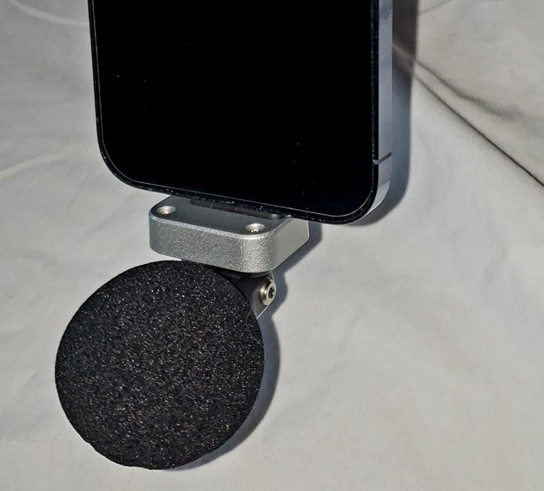 May include: A black smartphone attached to a silver metal bracket with a black, round, foam microphone. The phone is positioned above the microphone, which is connected to the bracket via a small hinge. The background is a light, neutral color.