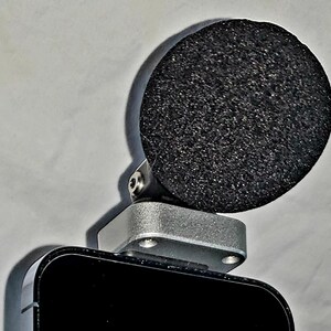 May include: A black microphone with a round, textured, black foam windscreen. The microphone is attached to a silver-colored metal base, which is mounted on a black rectangular device. The background is a plain, off-white surface.