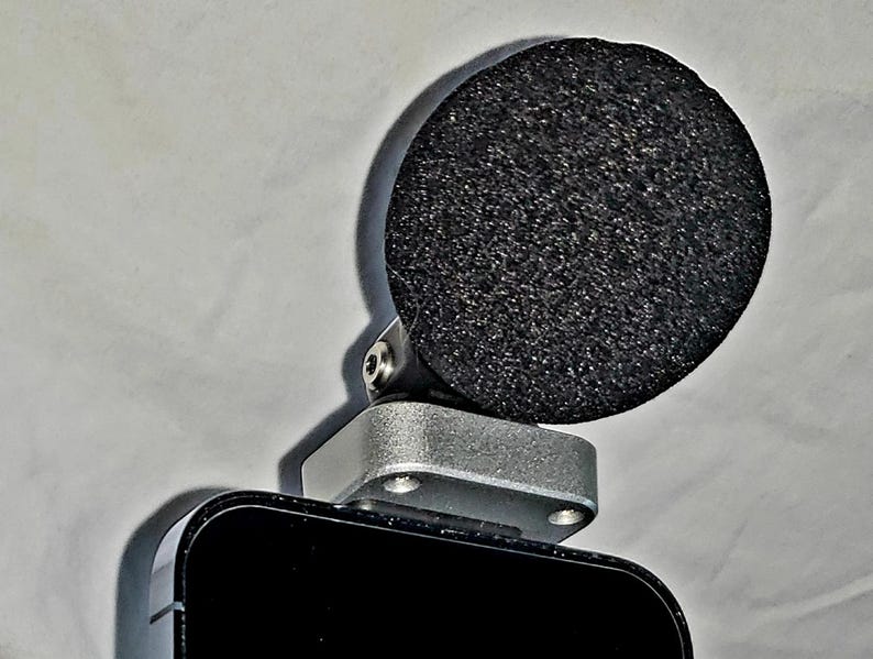 May include: A black microphone with a round, textured, black foam windscreen. The microphone is attached to a silver-colored metal base, which is mounted on a black rectangular device. The background is a plain, off-white surface.