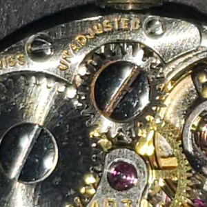 May include: Close-up of a watch mechanism with intricate gears, screws, and jewels. The words "UNADJUSTED" and "SWISS" are visible. The metal components have a silver and gold color.