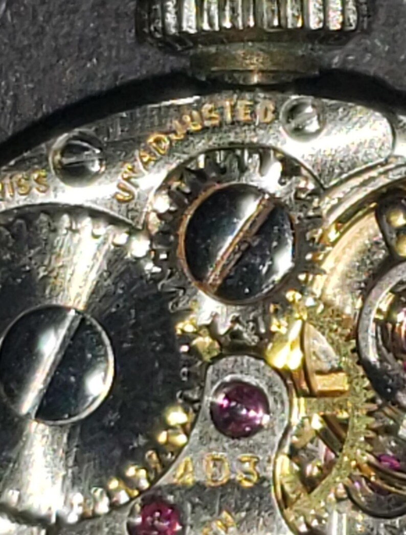 May include: Close-up of a watch mechanism with intricate gears, screws, and jewels. The words "UNADJUSTED" and "SWISS" are visible. The metal components have a silver and gold color.