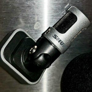 May include: A silver and black microphone with the brand name "SHU" printed on the side. The microphone is mounted on a black stand. A black foam cover is visible in the background. The microphone is designed for recording.
