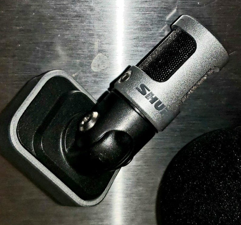 May include: A silver and black microphone with the brand name "SHU" printed on the side. The microphone is mounted on a black stand. A black foam cover is visible in the background. The microphone is designed for recording.