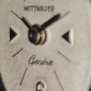 May include: Close-up of a vintage Wittnauer watch face. The watch face is off-white with black hands and markers. The word "WITTNAUER" is printed at the top, and "Gen&egrave;ve" is written in cursive below the center.