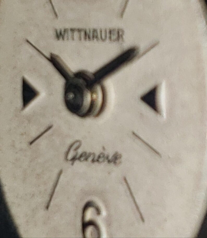 May include: Close-up of a vintage Wittnauer watch face. The watch face is off-white with black hands and markers. The word "WITTNAUER" is printed at the top, and "Gen&egrave;ve" is written in cursive below the center.