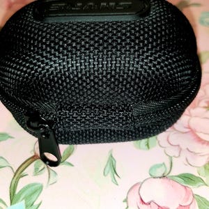 May include: Black, rounded carrying case with a zipper closure. The case has a textured, woven appearance and a black zipper. The top of the case has the brand name "SHURE" embossed in black. The case is on a floral background.
