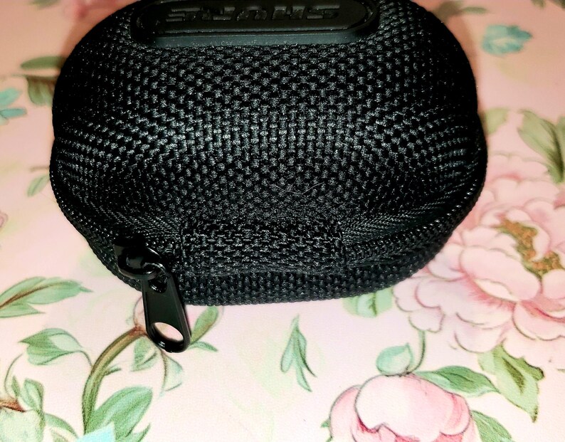 May include: Black, rounded carrying case with a zipper closure. The case has a textured, woven appearance and a black zipper. The top of the case has the brand name "SHURE" embossed in black. The case is on a floral background.