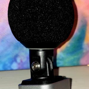 May include: A black microphone with a rounded, foam head. The microphone has a silver base and a black stem. The base has a lightning connector. The background is a colorful tie-dye pattern.