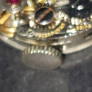 May include: Close-up of a watch mechanism with gold and silver components. The watch features a small, round crown and intricate internal gears. The word "SWISS" is visible on the watch face.