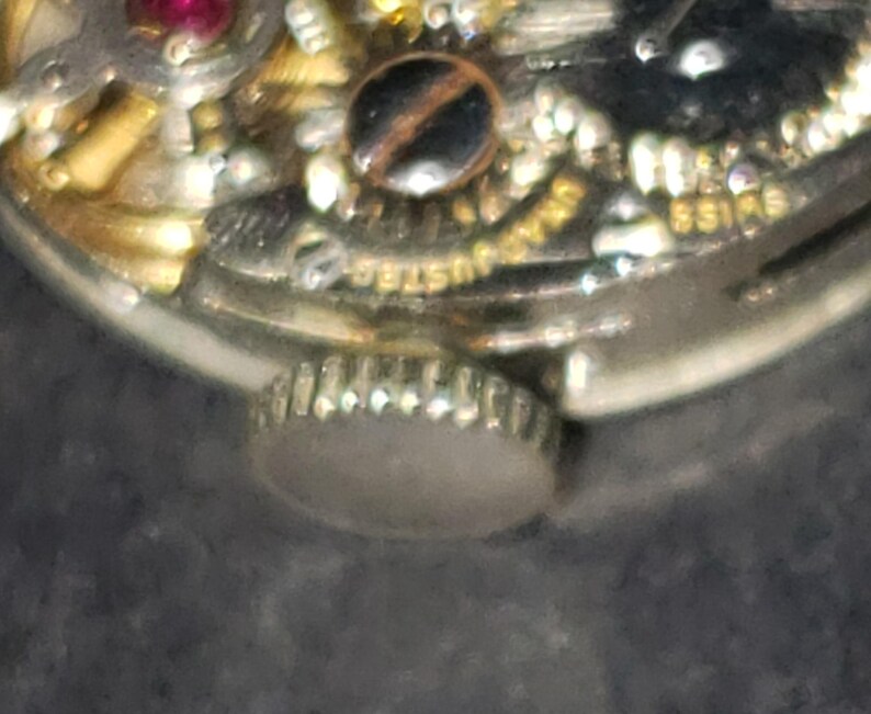 May include: Close-up of a watch mechanism with gold and silver components. The watch features a small, round crown and intricate internal gears. The word "SWISS" is visible on the watch face.