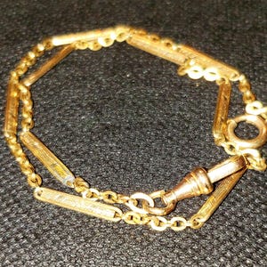 May include: A gold-colored watch chain with a series of rectangular and circular links. The chain has a clasp on one end and a ring on the other. The watch chain is displayed on a black surface.